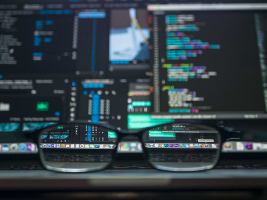 Fokussiert ist eine Brille die auf einem Tisch liegt. Im Hintergrund sieht man einen Computer mit hoher Rechenleistung.